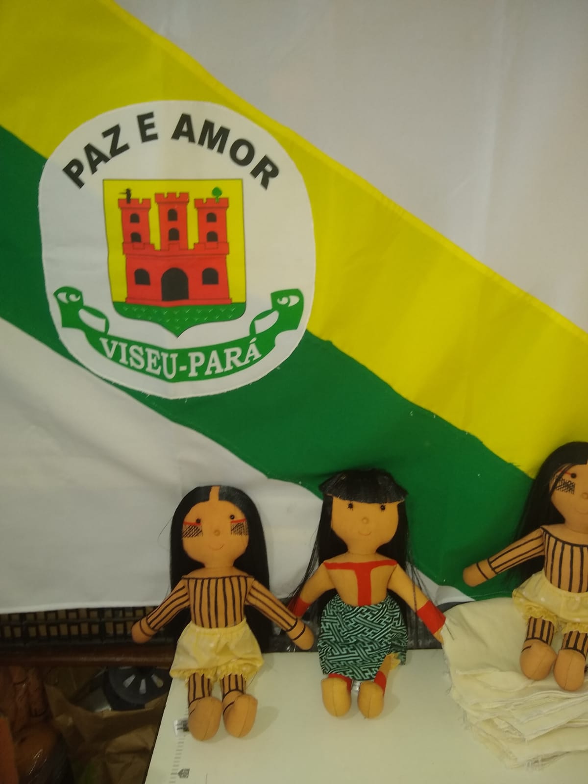 Essa bandeira é do Município de Viseu, Pará, cidade onde Luakam nasceu. As bonecas estão junto à bandeira porque elas dão muito orgulho para os conterrâneos. Uma foto muito importante para Luakam e seu povo, porque os conterrâneos sabem que Viseu está no mundo através das Bonecas Anaty.