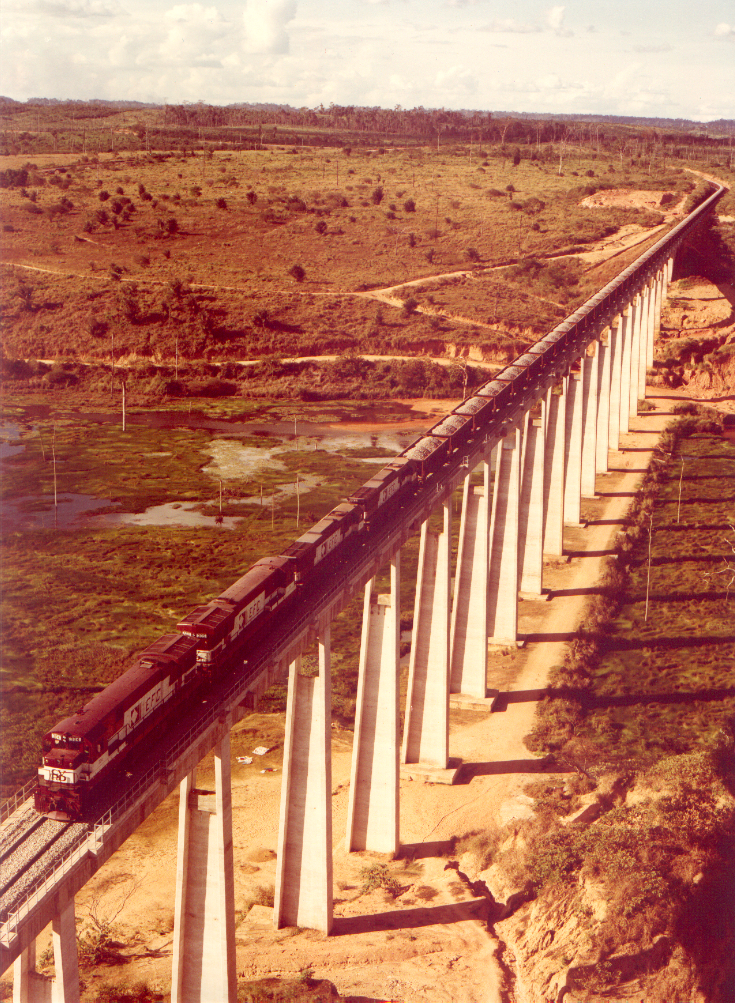 Locomotiva com vagões de brita em ponte ferroviária.