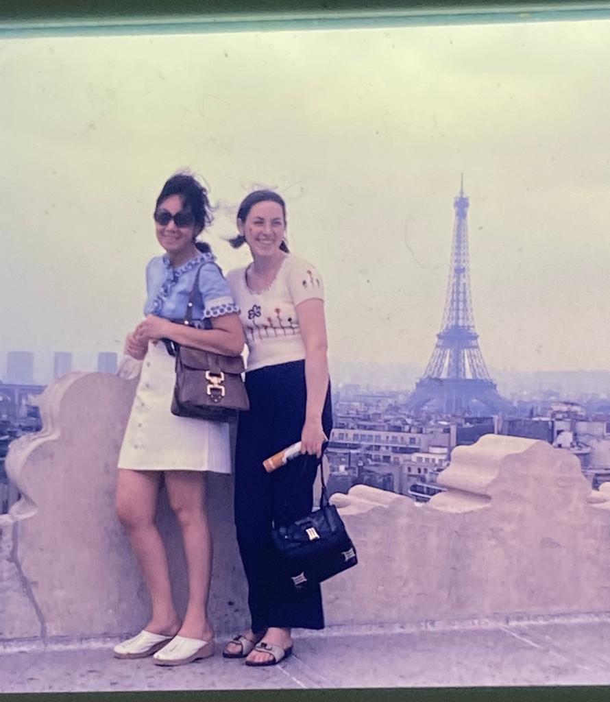 Débora Barenboim, vestida de camiseta clara e calça, posa para uma foto em Paris, ao lado de uma amiga, com a Torre Eiffel ao fundo.