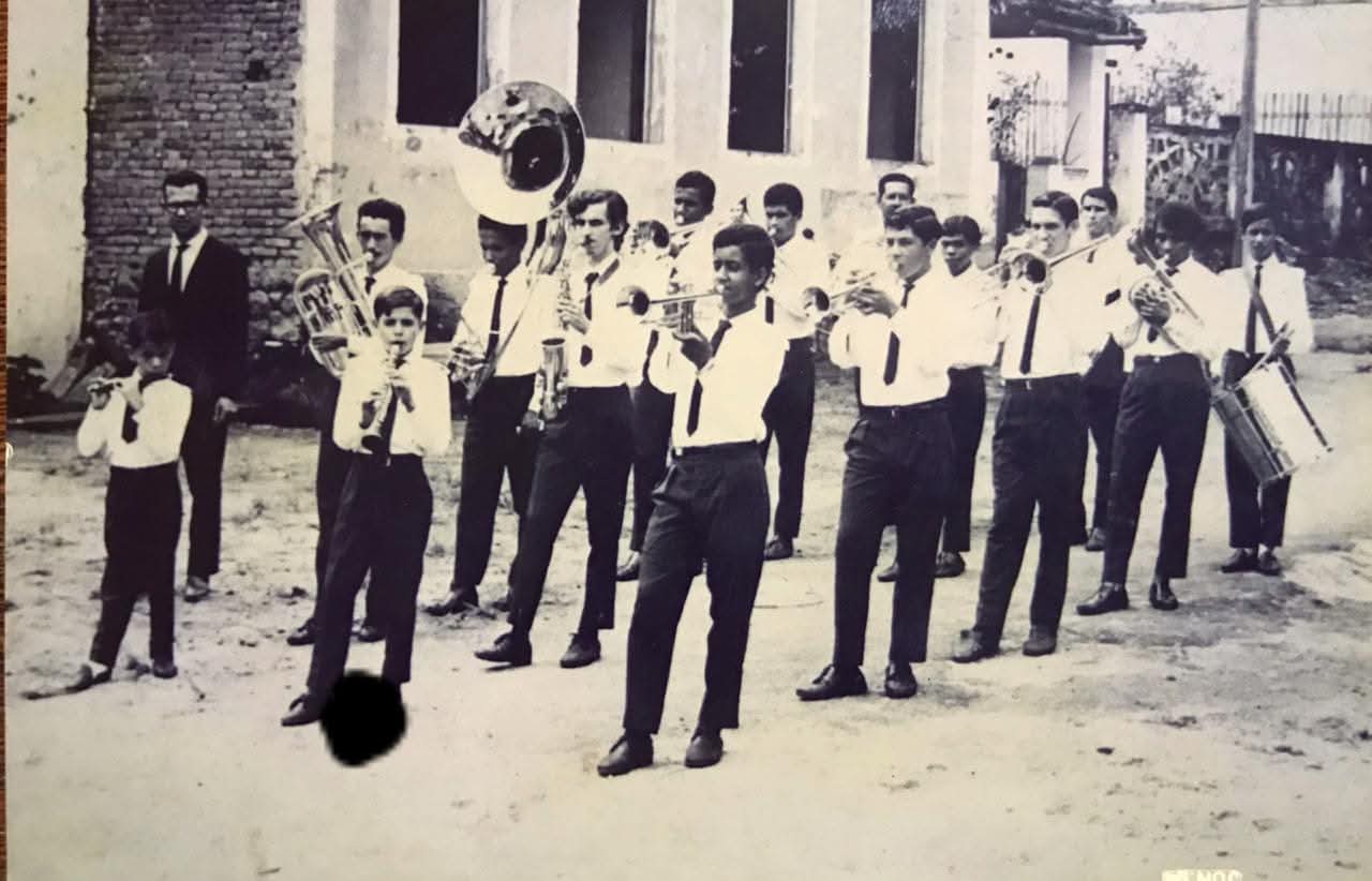 Registro histórico da Banda Lira Leopoldinense, tradicional banda musical de Santa Leopoldina, regida pelo saudoso maestro José Helena de Paula. A fotografia mostra os integrantes da banda durante uma apresentação pelas ruas da cidade, representando a forte tradição musical e cultural do município. Na imagem, Bebeto Pereira aparece tocando saxofone, participando ativamente das atividades musicais da banda na juventude.
