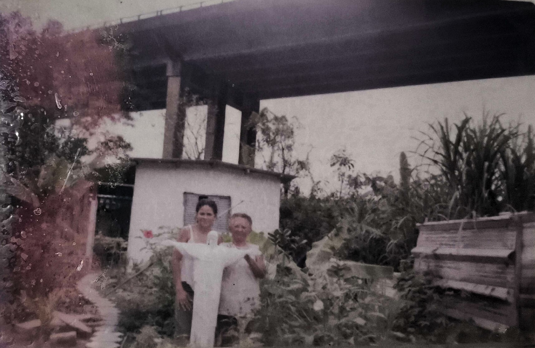 Adriana aparece posando ao lado da sogra, Dona Martinha, diante da imagem do Cristo que atualmente marca a entrada do Projeto AsMaras. O registro foi feito nos fundos do espaço da ONG De Bem com o Mangue, em Cubatão. A casa retratada corresponde ao local onde hoje vivem Adriana e seu marido, Sebastião, conhecido como Zumbi. Ao fundo, é possível identificar a Rodovia dos Imigrantes, compondo o cenário que conecta memória pessoal, território e comunidade.