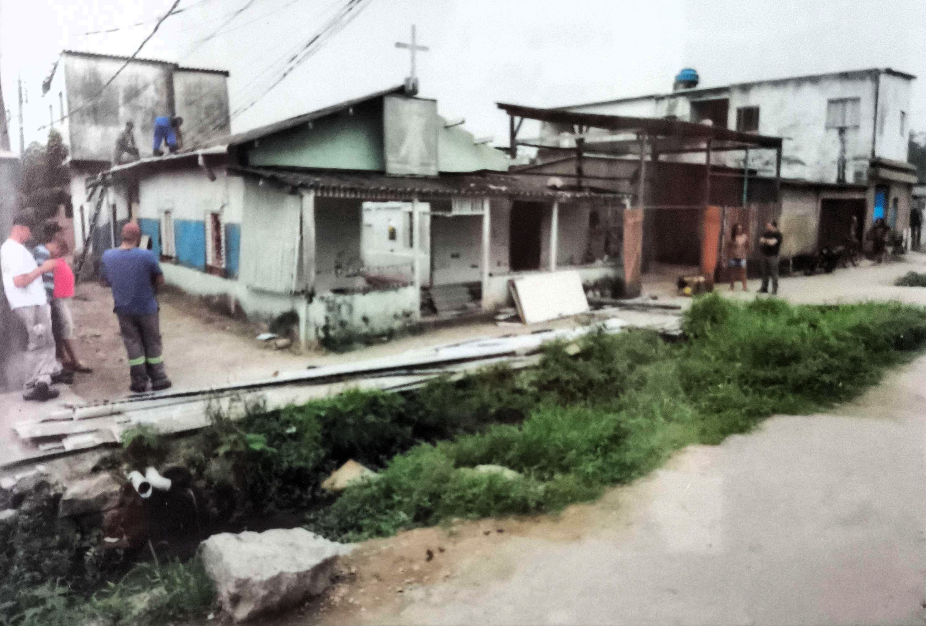 Nesta imagem, observamos a fachada da Igreja da Divina Misericórdia, no Ponto Final da Vila Esperança, com a cozinha de concreto ao fundo. A base da igreja já era de concreto, mas as paredes permaneciam em madeira. Em primeiro plano, aparece o córrego totalmente exposto, revelando as condições precárias da região e os desafios enfrentados pela comunidade para manter seu espaço de fé.