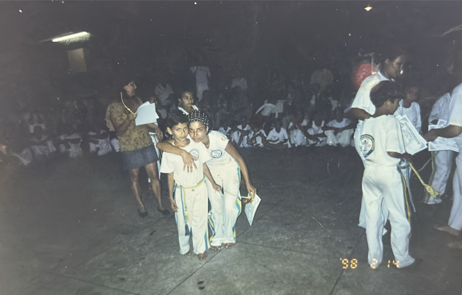 Registro do batizado de capoeira do Grupo Brasil/Suíça, no antigo Bingo Vegas, em Cubatão. Patrícia aparece ao lado de seu afilhado Meison, em um momento ritual de iniciação na capoeira. A fotografia também se relaciona à trajetória de Patrícia como capoeirista, marcada pela formação, pelo reconhecimento e pela conquista de diploma, reafirmando a capoeira como espaço de aprendizado, pertencimento e continuidade cultural.