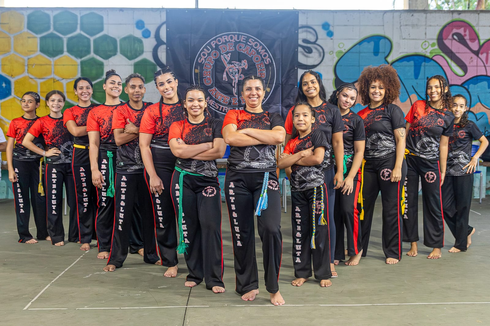 Patrícia aparece acompanhada de suas alunas durante um evento de capoeira. À sua esquerda estão Ana Clara, Isa, Letícia, Lívia, Paloma e Beatriz; à direita, Larissa, Ana, Nathália, Samira, Rachel, Mylla e Beatriz. A fotografia registra o grupo reunido, destacando a presença feminina na capoeira e a relação entre mestra e alunas em um contexto coletivo e formativo.