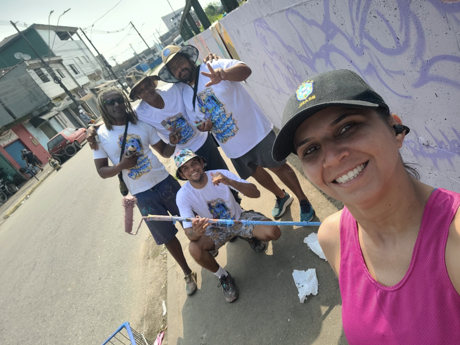 Patrícia aparece ao lado de integrantes do projeto da Rumo durante a ação de grafitar o muro da Vila Esperança. A imagem registra o artista Marco Tuim em atividade pelo Projeto da Linha Pra Frente, enquanto o grupo participa do processo de colorir e transformar a paisagem do bairro.