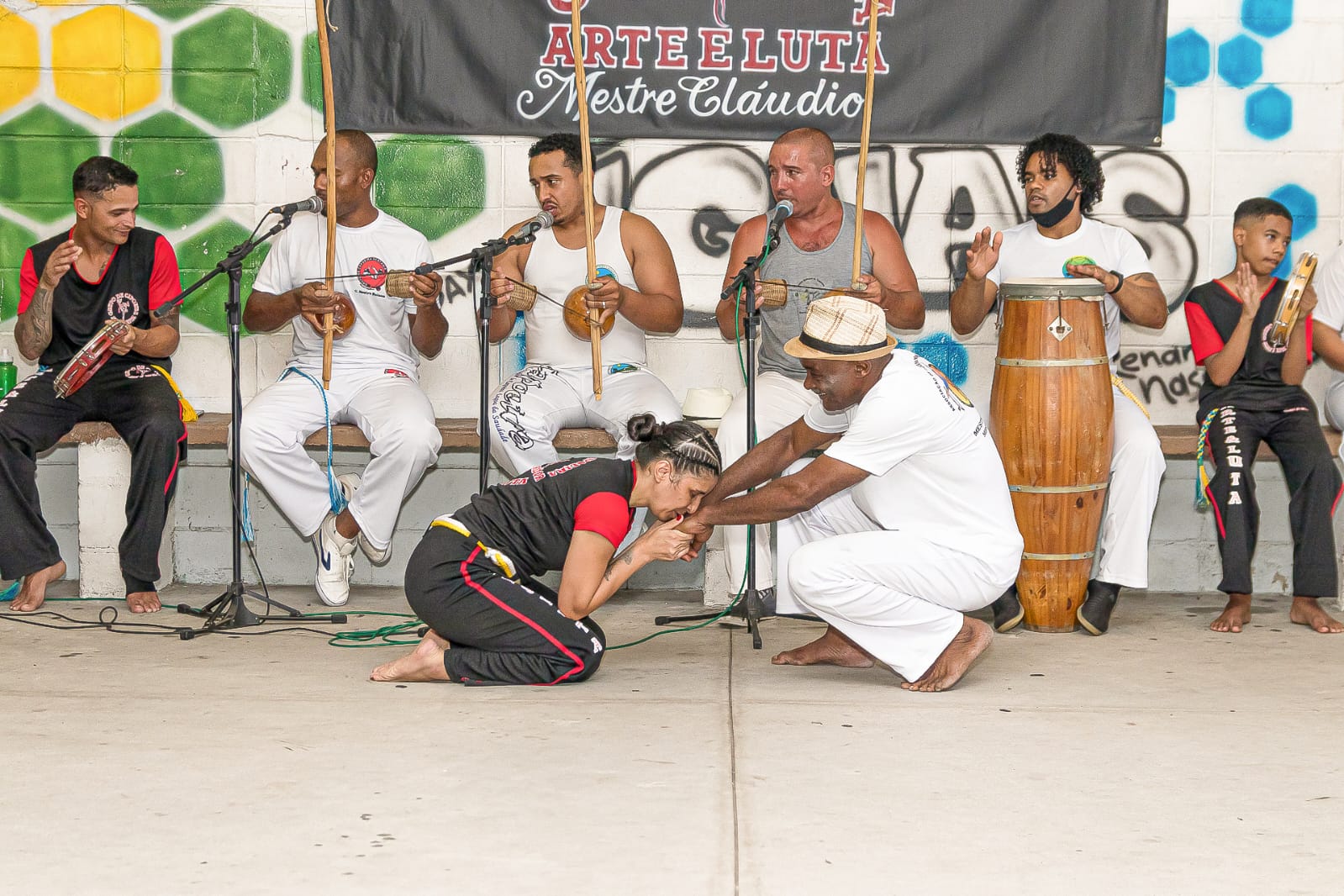 Na formatura da capoeira, Patrícia em gesto a Mestre Geraldo que simboliza respeito. A fotografia revela não apenas a relação entre mestre e discípulo, mas também o elo invisível que une gerações na capoeira. É um instante de honra, onde o aprendizado se transforma em reconhecimento e a roda se torna espaço de afeto e tradição.