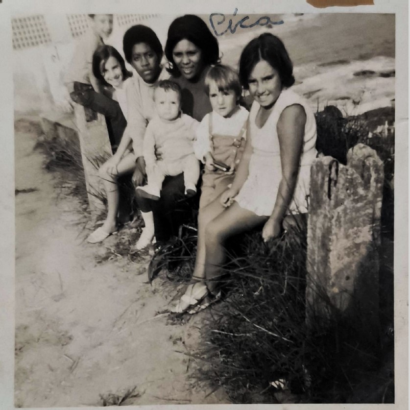 No centro da imagem está Dona Cícera, ainda jovem, em um momento de convivência com amigos e crianças, datado entre o final dos anos 1960 e início dos anos 1970. Ela aparece com um bebê no colo, cercada por outras crianças, em uma cena que transmite afeto e coletividade. O registro fotográfico, raro para o período e contexto social, representa não apenas um instante de encontro, mas também a valorização da memória e laços comunitários.