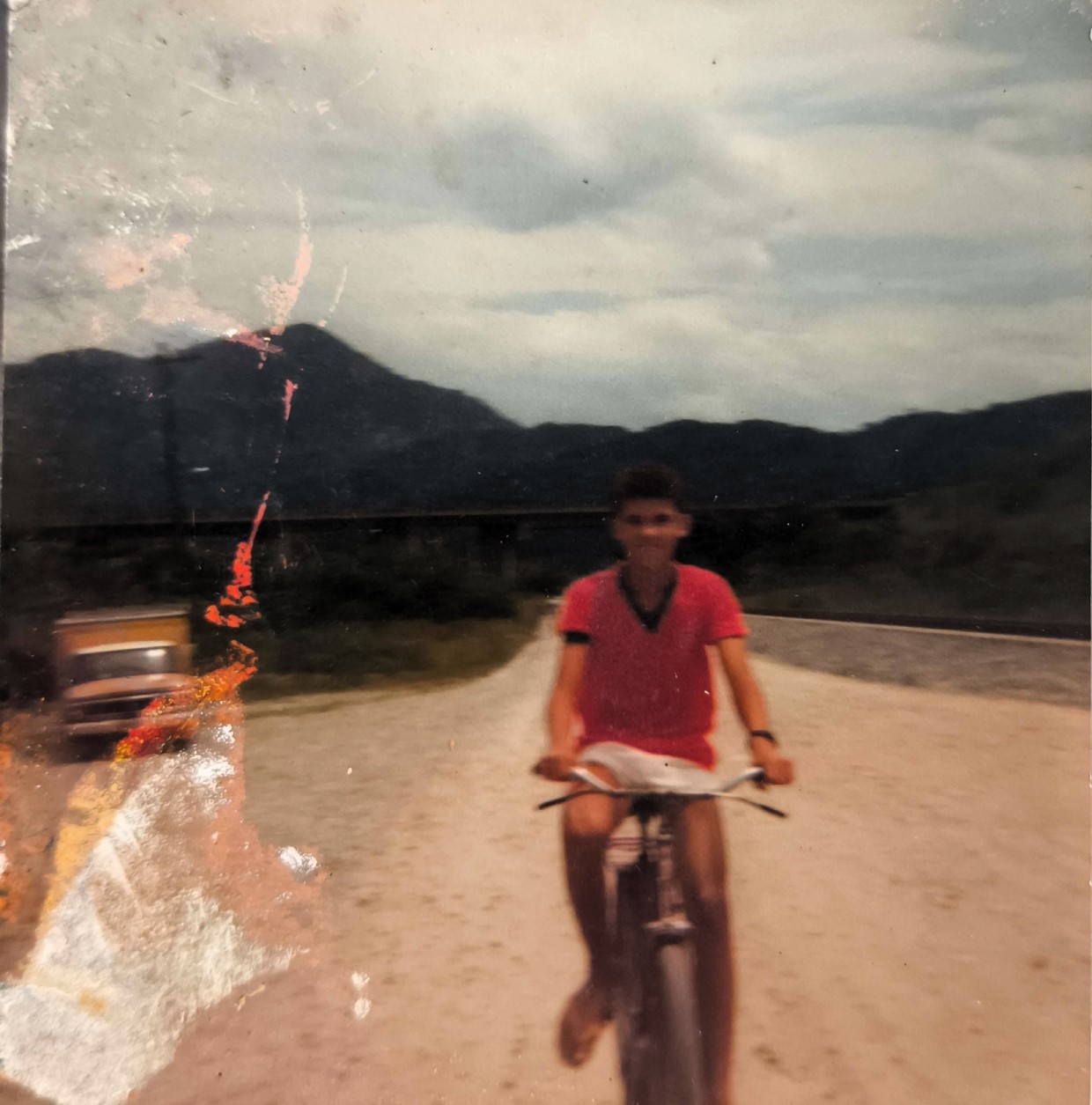 A imagem retrata Patrique, filho de Dona Cícera, ainda adolescente, sobre uma bicicleta, em registro feito na década de 1980. Ao fundo, observa-se a paisagem inóspita da Vila Esperança, em Cubatão, marcada por ruas de terra e amplos espaços vazios. Na época, a região apresentava características rurais, com poucas construções e grande distância entre as moradias. O cenário contrasta com o que viria a se tornar nas décadas seguintes: uma área densamente povoada e marcada pela intensa ocupação urbana. O registro carrega valor documental ao evidenciar a transformação territorial e social da comunidade.