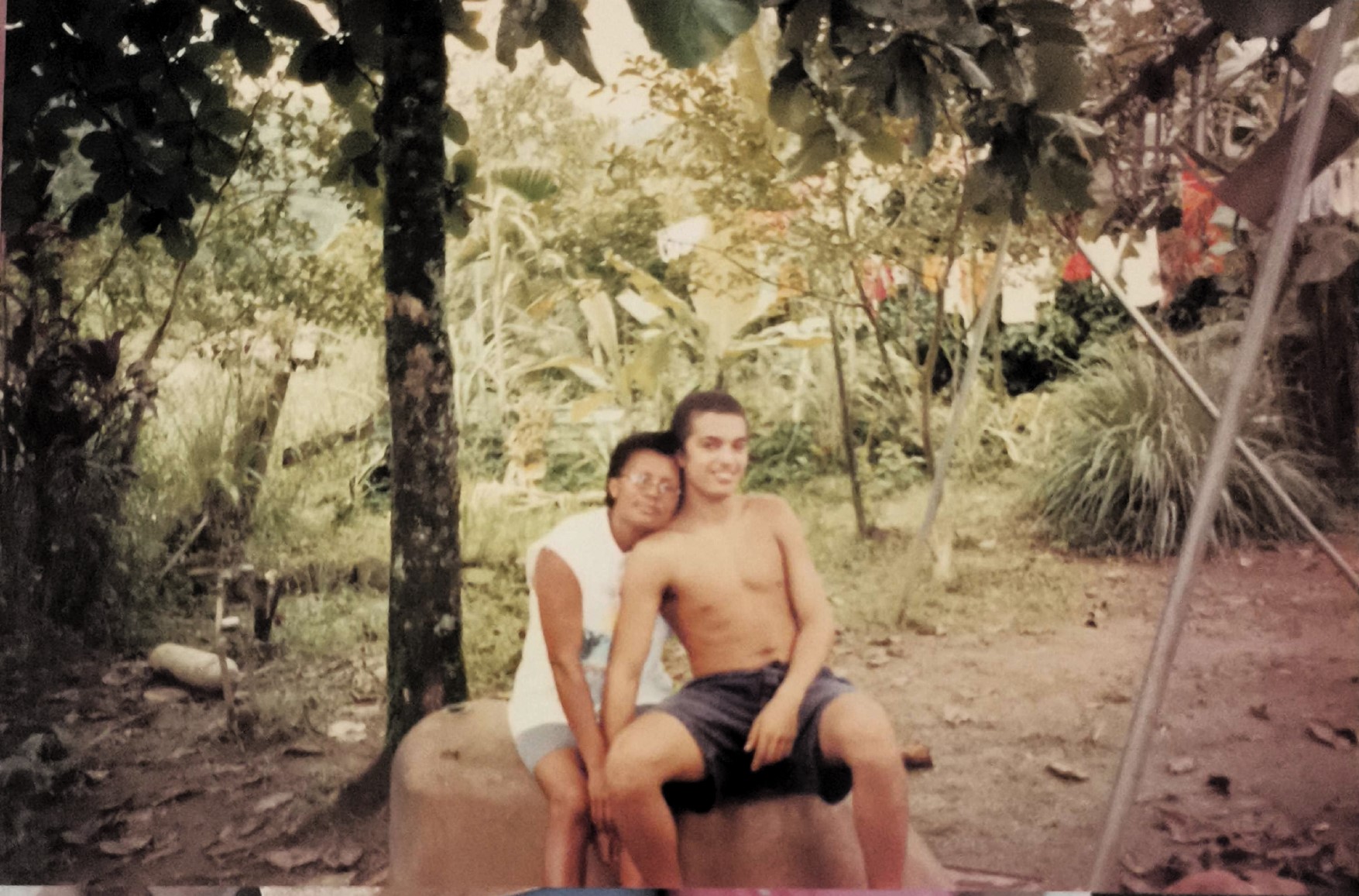 A imagem mostra Dona Cícera ao lado de seu filho Plínio, então adolescente, no quintal dos fundos de sua casa na Vila Esperança, em Cubatão. O registro revela não apenas o vínculo afetivo entre mãe e filho, mas também a paisagem da região no início de sua ocupação: uma área marcada por vegetação abundante e ausência de construções, com características próximas a uma zona rural. A imagem é um testemunho da vida cotidiana e da configuração territorial da comunidade em seus primeiros anos.