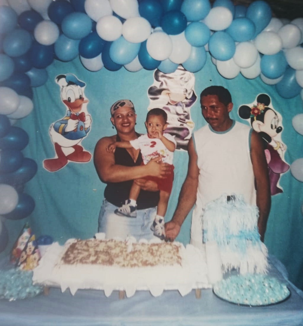 Celebração do aniversário de Artur, filho de Alfredo, conhecido como Cabeça, realizada na Vila Esperança. Atrás da mesa do bolo estão Alfredo, sua companheira Maria Valdete, paranaense, do interior de Curitiba, e o pequeno Artur. A fotografia retrata um momento íntimo e afetivo da vida familiar, revelando a importância das comemorações simples. Hoje adulto, Artur é lembrado pelo pai como um jovem dedicado aos estudos e ao trabalho.