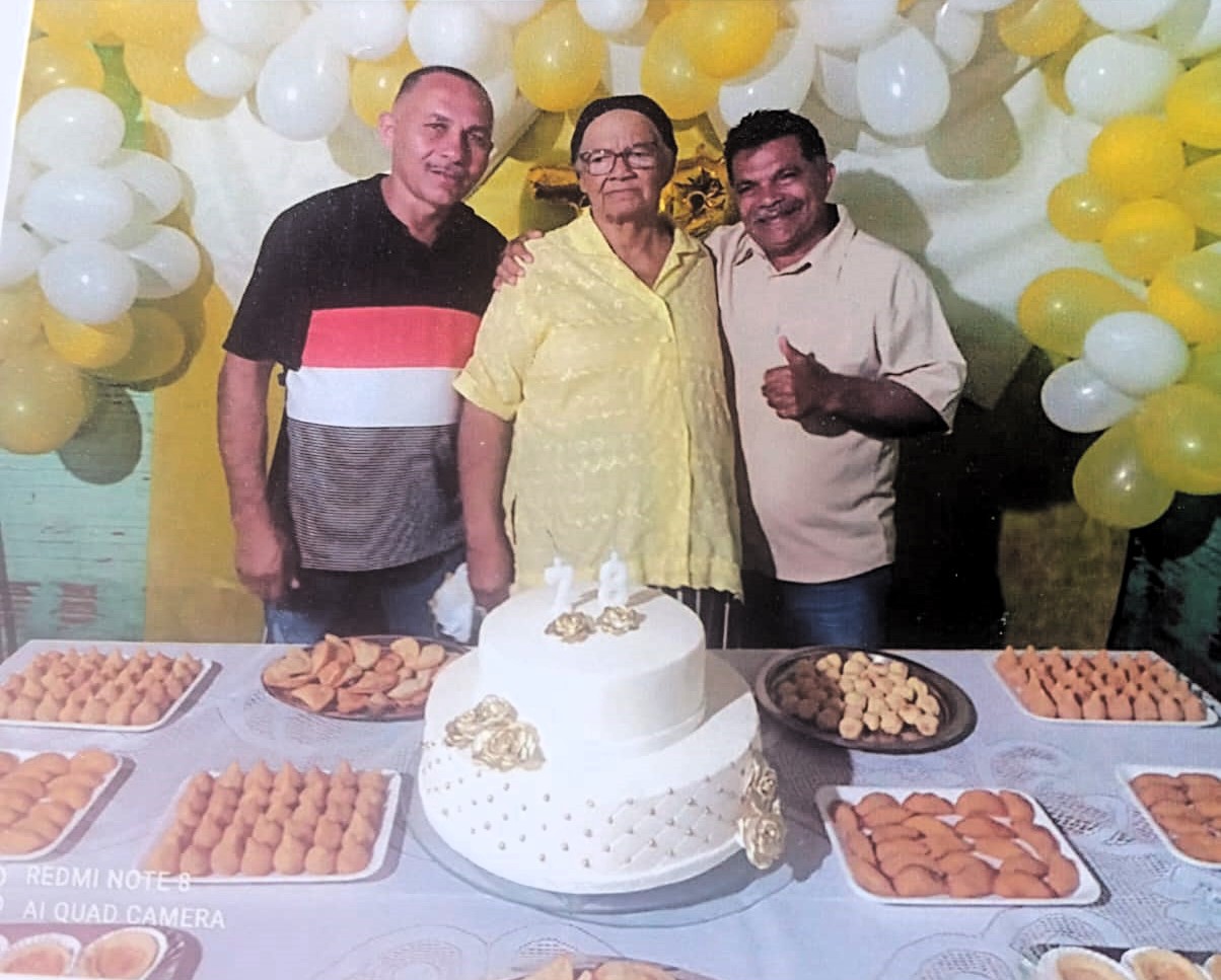 Fotografia da comemoração do aniversário de Dona Zefa, mãe de Alfredo, realizada no sítio Olho D’Água Cercado, onde ele nasceu e ela vive até hoje. A imagem celebra seus 78 anos de vida e destaca a relação de orgulho, respeito e admiração construída entre mãe e filho. Dona Zefa é lembrada como uma mulher forte, firme e cuidadora, figura central na formação familiar, reforçando a importância da matriarca como pilar afetivo e moral da família. A fotografia preserva a celebração da vida, da resistência e da ancestralidade.