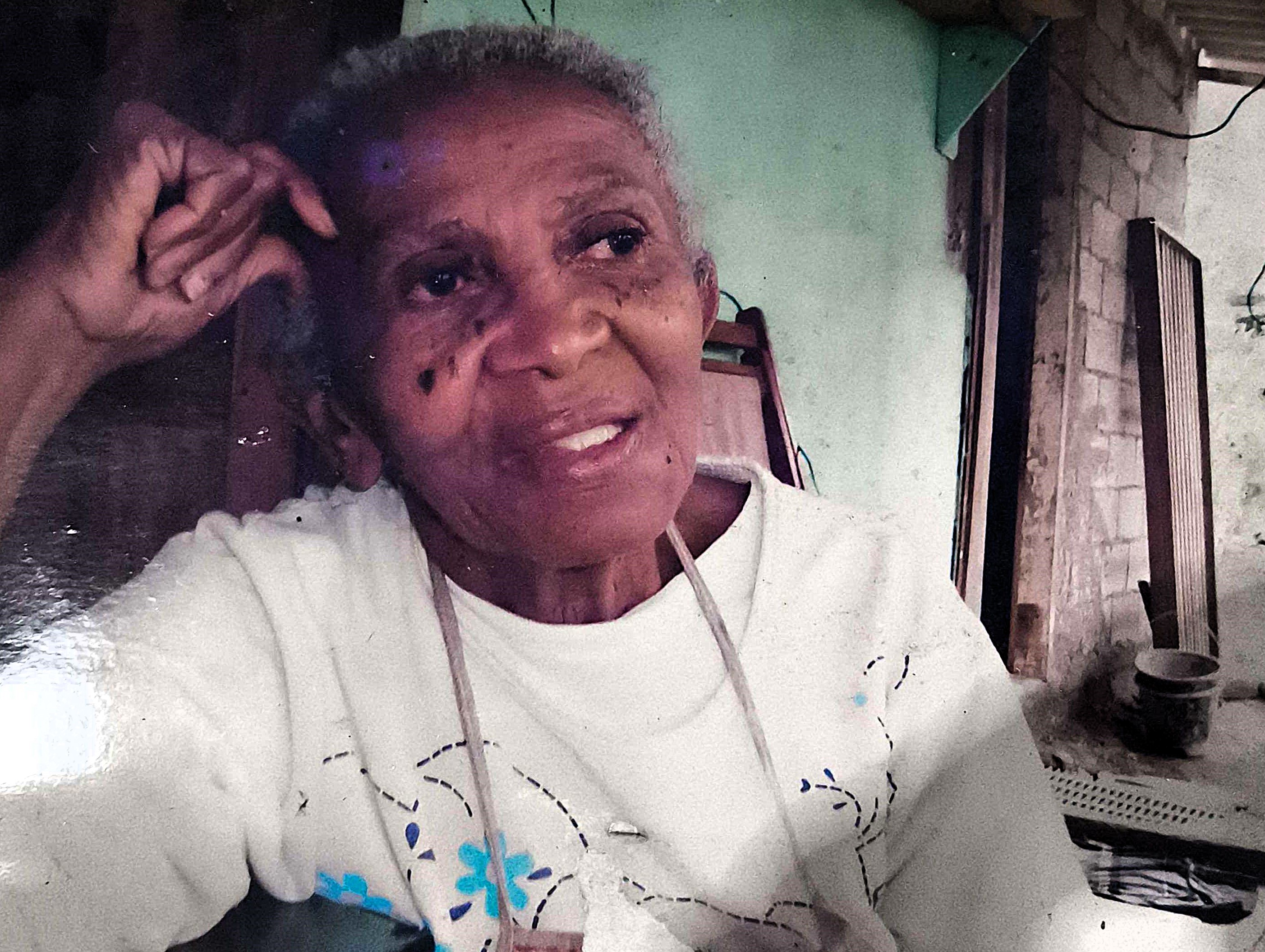 A imagem retrata Dona Martinha, mãe de Zumbi, em um momento introspectivo. Moradora da Ilha Bela desde o início dos anos 1980, ela foi reconhecida como uma importante líder comunitária e como forte inspiração para o trabalho de seu filho, Sebastião, o Zumbi, que segue atuante como liderança local. A fotografia valoriza a presença feminina nas lutas do território e a influência das mulheres na construção da vida comunitária.