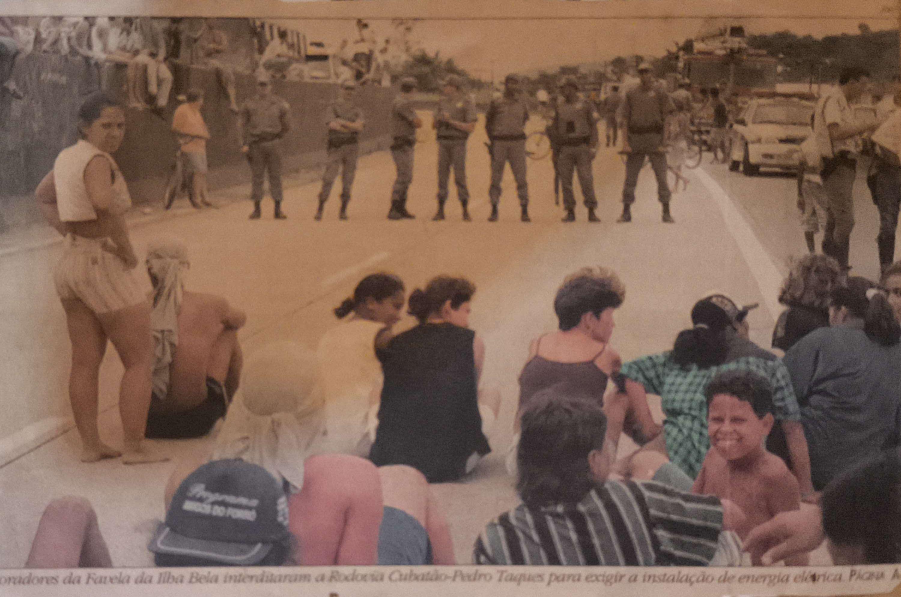 A fotografia registra a paralisação da Rodovia Padre Manoel da Nóbrega, na época chamada de Pedro Taques, organizada por lideranças comunitárias, entre elas Zumbi, como forma de reivindicar a instalação de energia elétrica na Vila Esperança. Moradores sentam-se na via principal enquanto enfrentam a presença da segurança pública. O ato evidencia a força coletiva da comunidade em defesa de condições dignas de vida.