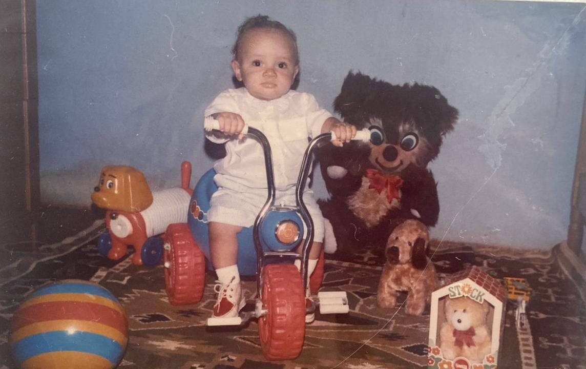 Hélcio em cima de uma motoca numa fotografia presente no álbum comemorativo de seis meses de idade, em outubro de 1985, na cidade de Itamaraju, BA.