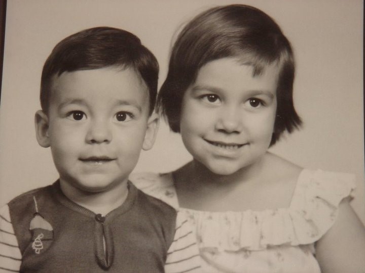 Guilherme e sua irmã, Carol, durante a infância, em uma sessão de fotografias em estúdio. A fotografia é um pôster e está pendurada na sala da casa da sala da mãe de Guilherme. Ele relembra que a camisa de lã que vestia tinha um fio solto no pescoço que sempre o incomodava, é uma das primeiras memórias de sua infância.