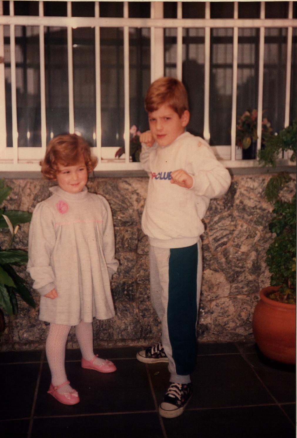 Milena tinha quatro ou cinco anos quando a fotografia aconteceu. Ao lado do irmão, Fábio, foi preparada pela mãe como um pequeno boneco para um evento no centro espírita que ela frequentava. Vestir a calça foi uma batalha. Antes do clique, houve choro. Houve recusa. Milena não queria ser fotografada. Entre tentativas frustradas e imagens que não sobreviveram ao tempo, o momento parecia perdido. Até que o irmão fez uma graça. Um sorriso mínimo escapou, quase por descuido, e foi ali que a câmera encontrou o que procurava. A foto que ficou não é a do choro, mas a do intervalo: esse breve acordo entre a vontade da mãe e o gesto espontâneo da filha. Um riso capturado no exato instante em que tudo quase falhou.