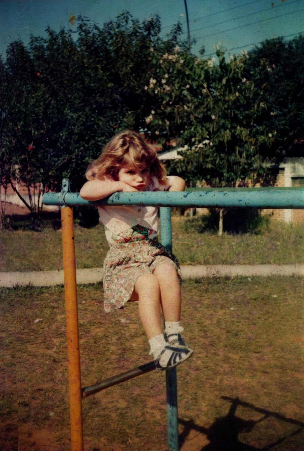 Aos cinco anos, Milena encara a câmera com uma expressão firme, quase brava, mas atravessada por um gesto que parece ensaiado. Há algo de teatral nesse rosto infantil, uma performance precoce, já anunciando uma personalidade que se constrói entre a pose e o jogo. A fotografia foi feita no Jardim Veleiros, na zona sul de São Paulo, bairro marcado pelo convívio intenso entre crianças e pela ocupação coletiva dos espaços. Nos fins de semana, a creche onde o irmão estudava se abria como território de brincadeira. Mesmo ainda pequena demais para ter vaga, Milena se sentia parte da escola: entre outras crianças, inventava pertencimentos. A imagem guarda esse limiar entre estar fora e dentro, entre brincar e representar, e revela um bairro que educa tanto quanto a instituição.