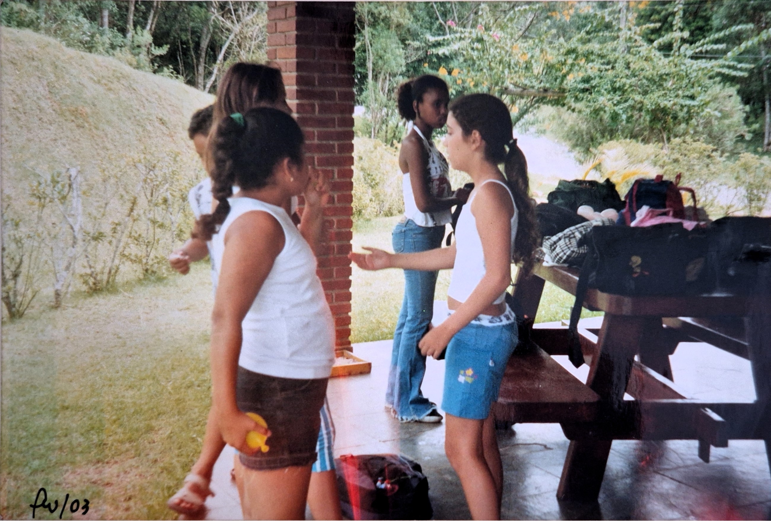 Aos doze anos, Milena está em uma chácara em Cotia, reunida com um grupo de teatro que atravessava os limites do bairro. Embora os encontros acontecessem na zona sul de São Paulo, perto de sua casa, o grupo misturava gente de dentro e de fora, ampliando amizades e horizontes. Na mão de Carina, amiga do bairro, está o gesto cúmplice de quem conhece bem a arte da molecagem, aquela malandragem leve que também atingia quem não estava jogando. Mais do que um dia de diversão, essa imagem marca um período de formação. No teatro, Milena encontrou algo que a escola não oferecia: o fortalecimento do grupo, a criação compartilhada, a ocupação da rua, as viagens, o prazer de inventar. Foi ali o primeiro contato com o mundo das artes, com figurinos, personagens e modos de existir outros. A lembrança de Teresa, assistente da professora, de cabelo rosa e estilo radicalmente distinto, permanece como sinal de ruptura e descoberta. A fotografia guarda o início desse deslocamento: entre bexigas d’água e risadas, o ensaio de uma sensibilidade que começava a se afirmar.