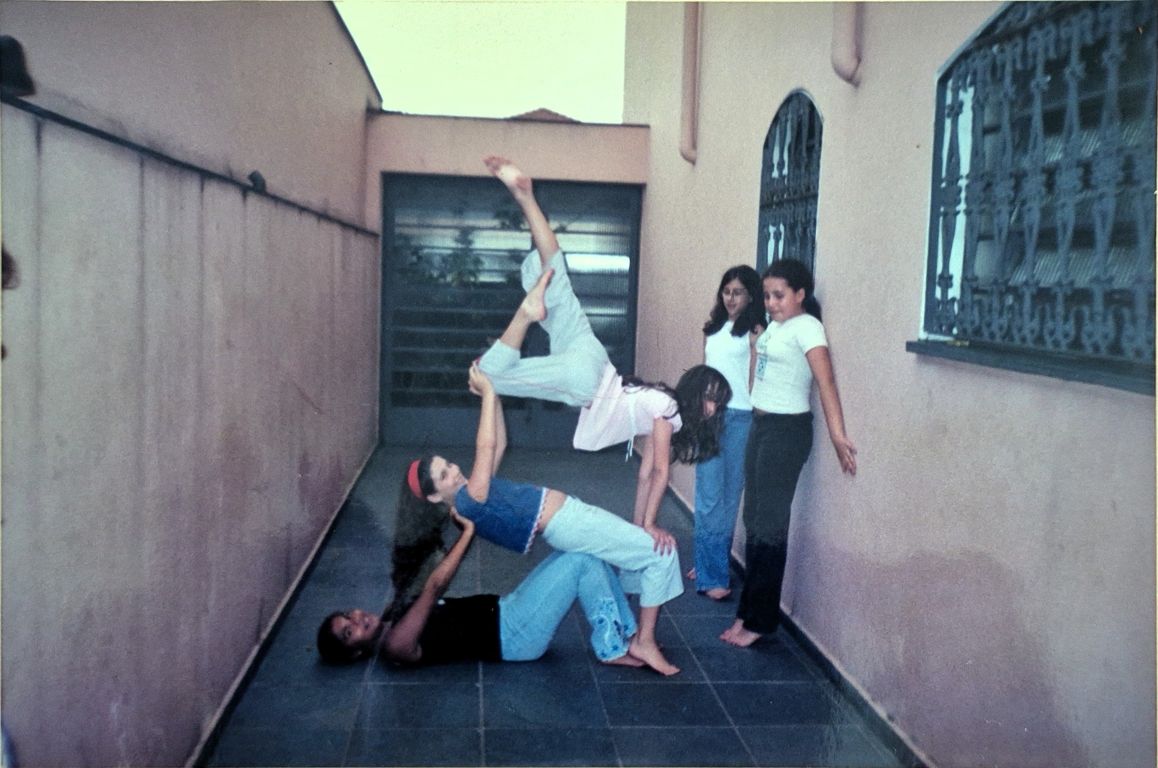 A fotografia registra um momento de liberdade e experimentação vivido no aniversário de 14 anos de Milena, no quintal de sua casa, na zona sul. Sem a presença direta de adultos, um grupo de meninas, amigas da escola e do teatro, entre risos, música e tentativas, elas encenam movimentos inspirados na ginástica artística, aprendidos com Natália, amiga e referência naquele instante, que praticava ginástica olímpica. O corpo coletivo se organiza em camadas de apoio e confiança: Milena no chão sustenta Natália, enquanto Jéssica lhe dá base. Outras duas meninas, a outra Jéssica e a Carol, permanecem observando, sem saber exatamente como participar. O gesto é lúdico, colaborativo e inventivo, mais próximo do teatro do que do esporte formal. A imagem revela uma experiência profundamente feminina, marcada pela ausência de vigilância, pela autonomia concedida pela mãe e pela ocupação livre do quintal como espaço de criação. O ato de “apresentar”, mesmo sem público definido, transforma o cotidiano em performance. A fotografia é feita pela mãe, que, ao observar a cena, decide preservá-la, reconhecendo a potência daquele instante de amizade e descoberta do próprio corpo.