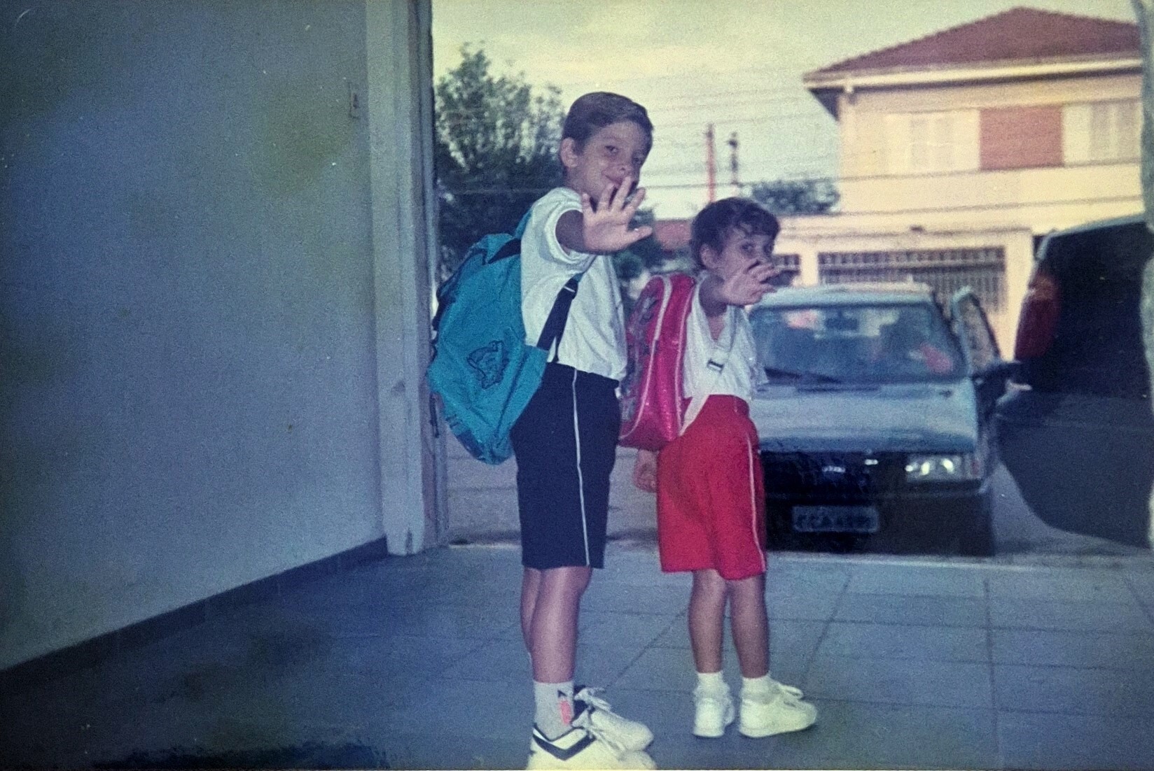 A fotografia registra o primeiro dia de Milena na primeira série, diante da escola estadual do bairro, a “escola grande”, já conhecida por meio do irmão mais velho. A imagem guarda a tensão silenciosa de um começo: o medo do desconhecido. Naquela manhã, ela sai cedo de casa ao lado do irmão Fábio. Excepcionalmente, o pai oferece carona, quebrando a rotina das longas caminhadas de cerca de vinte minutos, comuns em um bairro sem transporte público. Mais do que marcar a entrada na vida escolar, a fotografia preserva a paisagem da rua central, ponto de passagem e de encontros. É ali que a infância se estende para além dos limites da casa. A rua funciona como eixo do bairro, lugar onde todos acabam se cruzando, e sua presença na imagem amplia o valor documental da cena. O gesto capturado é simples e cuidadosamente conduzido pela mãe, que fotografa e orienta: um “tchau” sincronizado, ensaiado, quase coreografado. A memória evoca o gosto inicial pelos estudos, o vínculo afetivo com a primeira professora, Alessandra, lembrada como a mais querida, e o percurso escolar que, mais tarde, encontraria no teatro um espaço de liberdade e invenção.