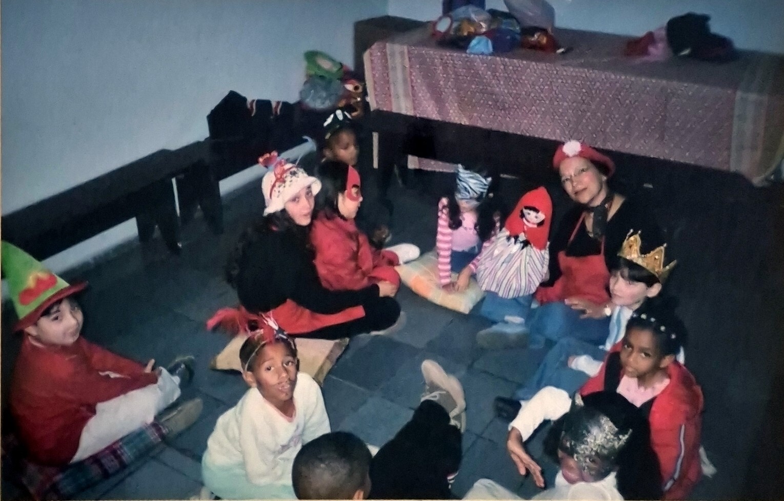 A fotografia registra um momento vivido no Neuchatel ou Centro Espírita Casa de Fabiano, no Jardim Veleiros, na Zona Sul, espaço que teve papel central na infância da Milena. O centro espírita funcionava como lugar de convivência, aprendizado e cuidado coletivo. Ali, as crianças permaneciam enquanto as mães participavam de palestras e sessões, ocupando um espaço destinado aos menores. Desde muito cedo, Milena passa a dividir a responsabilidade pelas crianças pequenas, inventando histórias, encenando pequenas peças de teatro e acolhendo bebês no colo. O centro também era um polo de atividades culturais, como aulas de flauta, violão, coral e teatro, além das ações sociais, com distribuição de sopa, roupas, remédios e brinquedos em datas comemorativas. A fotografia foi feita em um desses dias especiais. O contexto social do lugar atravessa a imagem de maneira silenciosa: Milena e sua família não se reconheciam plenamente nem como beneficiários, nem como doadores. Habitavam um espaço intermediário, marcado por necessidades reais, mas também por uma casa relativamente estável e bem localizada.