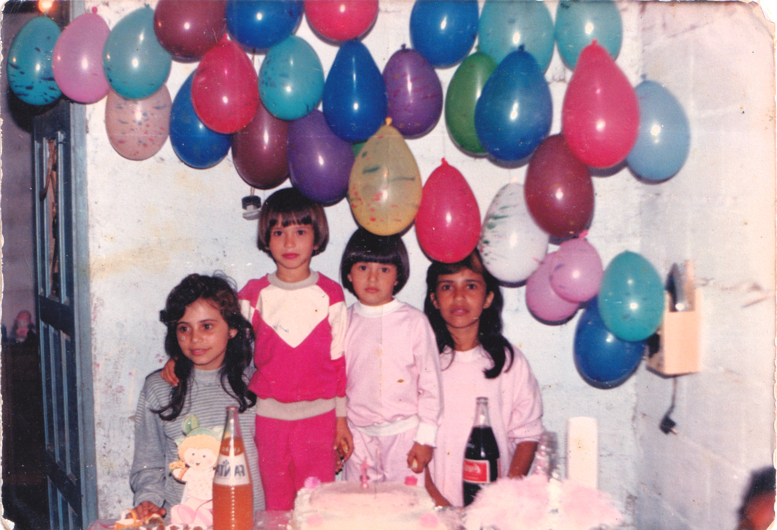 Na imagem aparecem Sandra, Juliana, Sabrina e Fabiana, em um dia marcante da infância: o aniversário de seis anos da Juliana. Foi nessa ocasião que Sandra, então com cerca de doze anos, cortou o cabelo da Juliana e da Sabrina em uma franjinha “tigelinha”, usando a tesoura que a mãe havia lhe entregue. A Sabrina gostou do resultado, achou o corte bonito e bem redondinho, enquanto a Juliana não ficou tão satisfeita. A comemoração aconteceu no Parque Santo Antônio, na casa do pai, ainda muito simples: ao fundo, vê-se o quarto pintado, sem reboco, com telhado de telha aparente. As festas eram modestas, com um bolo simples e alguns salgadinhos, mas carregadas de significado. Fotografias eram raras naquela época, já que a família não possuía câmera, um item caro para a realidade vivida. Não se sabe ao certo quem fez o registro, mas naquele aniversário houve algumas fotos, possivelmente feitas por algum amigo da família.