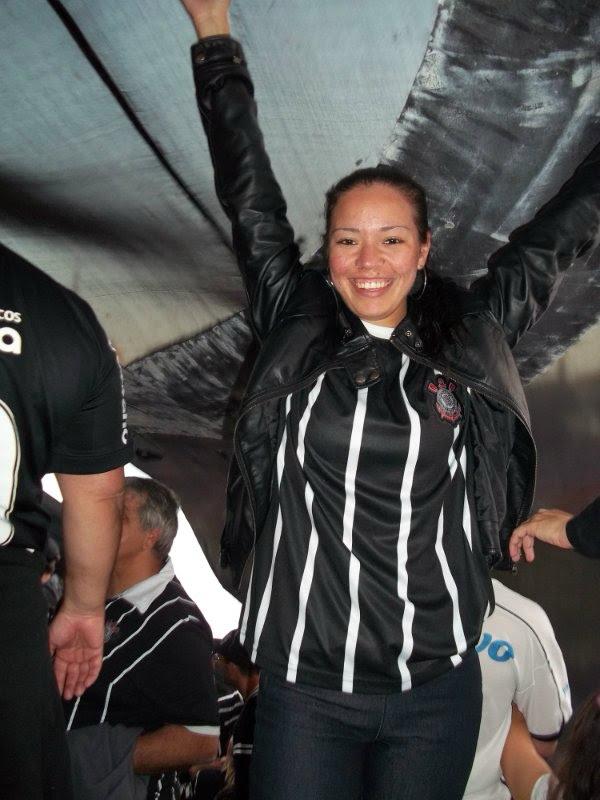 Primeira experiência de Dayana embaixo de um bandeirão, no Estádio do Pacaembu vestindo a camiseta do Corinthians. A imagem registra pertencimento, paixão esportiva e identidade coletiva.