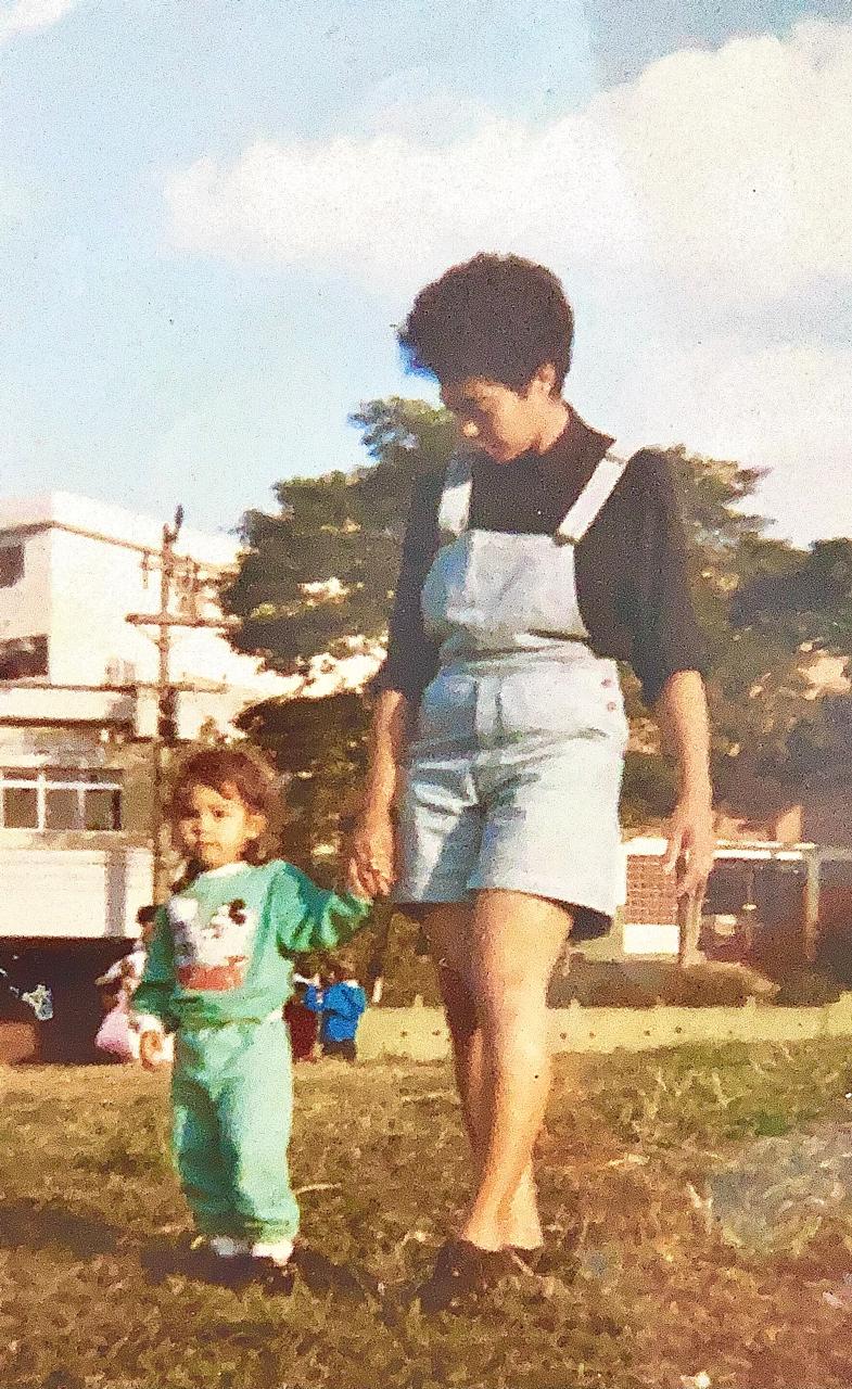 Dayana caminha de mãos dadas com sua mãe, Juju, em uma praça da cidade. A imagem revela cuidado, proteção e os primeiros passos guiados pelo afeto materno.