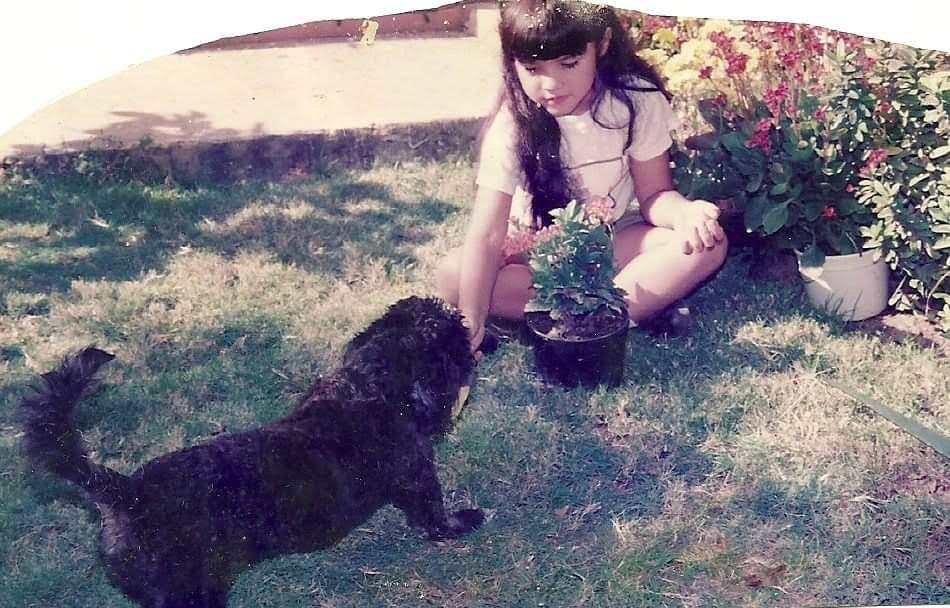Oriana aparece ao lado de Muñeca, sua primeira cachorra, em frente à casa de Cabimas onde viveu os primeiros cinco anos de sua vida. Não era um dia especial, mas tornou-se um momento valioso: recorda-se de que compraram flores ali, gesto simples que se transformou em hábito e prazer que a acompanha até hoje. A fotografia guarda a ternura da infância, a presença de Muñeca e o encanto das flores.