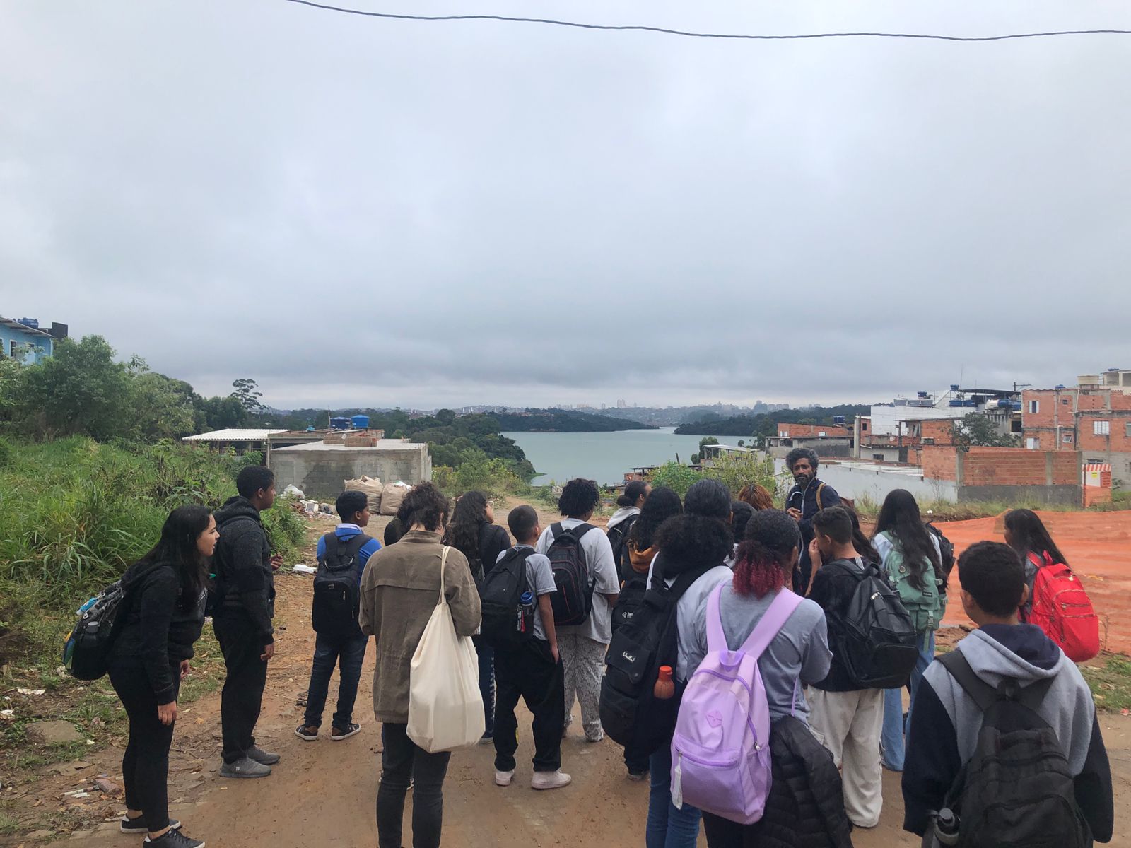 Estudantes e educadores participando de um percurso educador na região do Grajaú, em uma ação de docência compartilhada entre o Coletivo Imargem e escolas públicas da comunidade. Ao fundo, observa-se o contraste entre a ocupação urbana periférica e as águas da represa Billings sob um céu nublado.