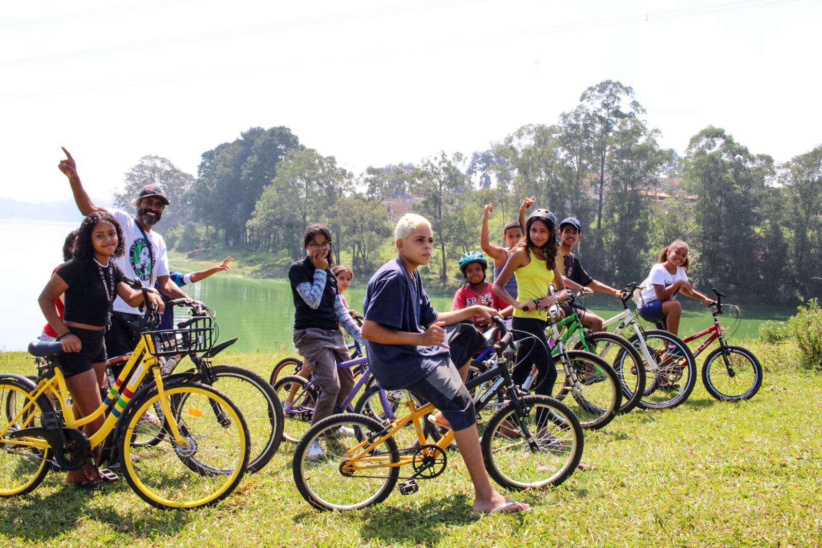 Crianças e adolescentes do Jardim Gaivotas se reúnem com suas bicicletas na margem gramada da represa Billings durante uma atividade do projeto Bikes Marginais. Coordenado pelo Coletivo Imargem, o percurso une lazer, mobilidade e educação ambiental, transformando as pedaladas em uma forma de explorar, reconhecer e valorizar a paisagem natural do Grajaú.