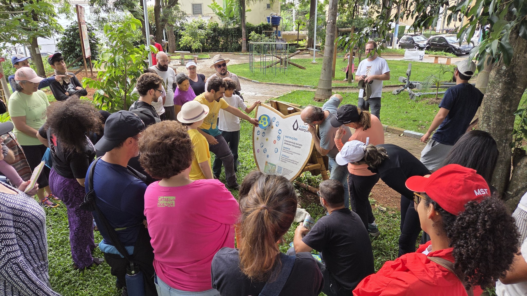 Alunos do SESC reunidos ao redor de um grande painel no Parque da Joia, acompanhando a explicação sobre o funcionamento do jogo de percurso criado para o espaço.