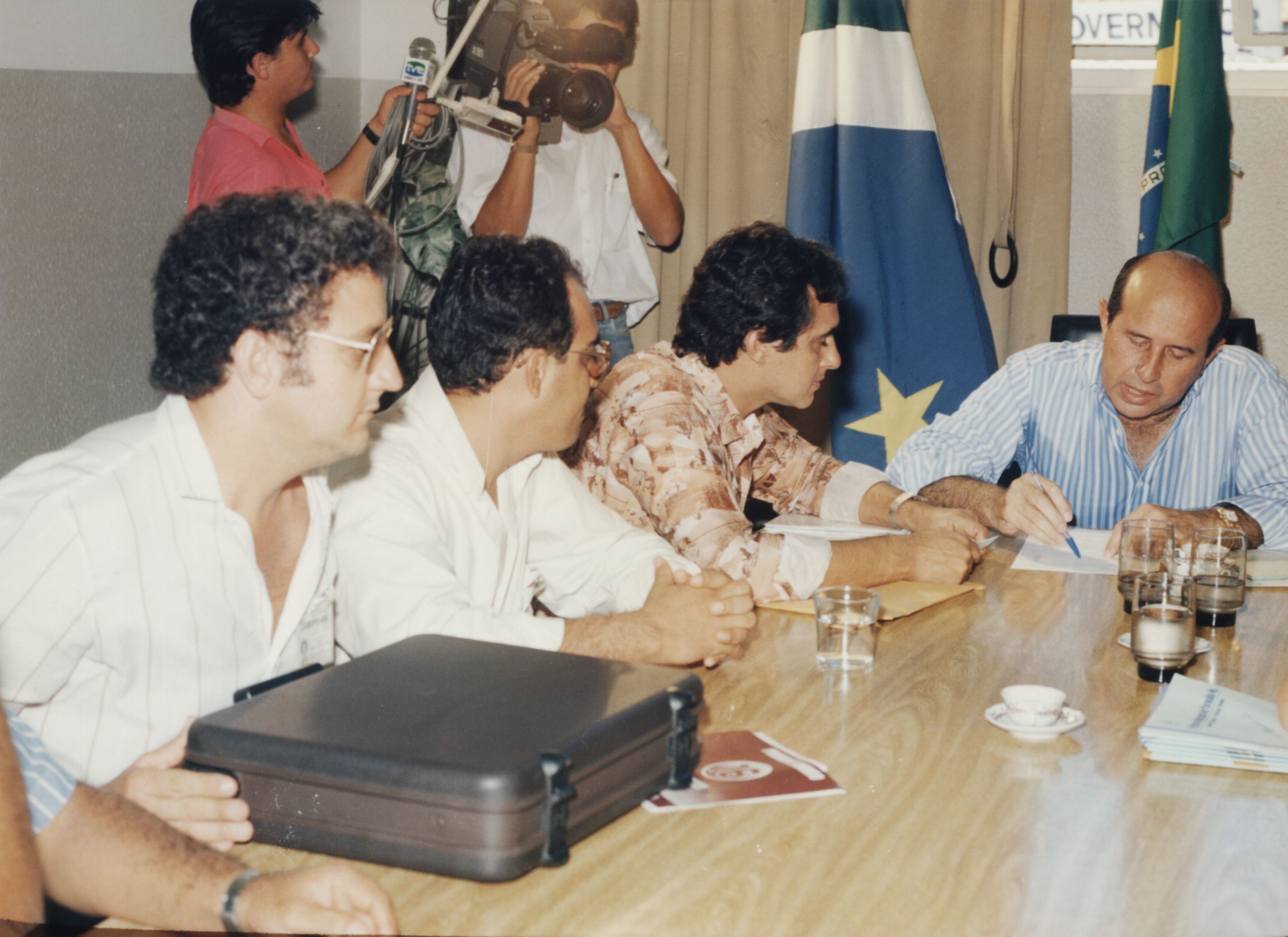 Da esquerda para a direita estão, Evauri, Marconis F. Alves (gerente regional da CTBC/Iturama), Wilson R. M. de Oliveira (prefeito de Paranaíba) e Marcelo Miranda Soares (governador do Mato Grosso do Sul) em audiência para tratar de assuntos sobre telefonia em Paranaíba. O governador assina documentos na presença dos outros, também sentados à mesa. Um repórter e um cameraman então em segundo plano.