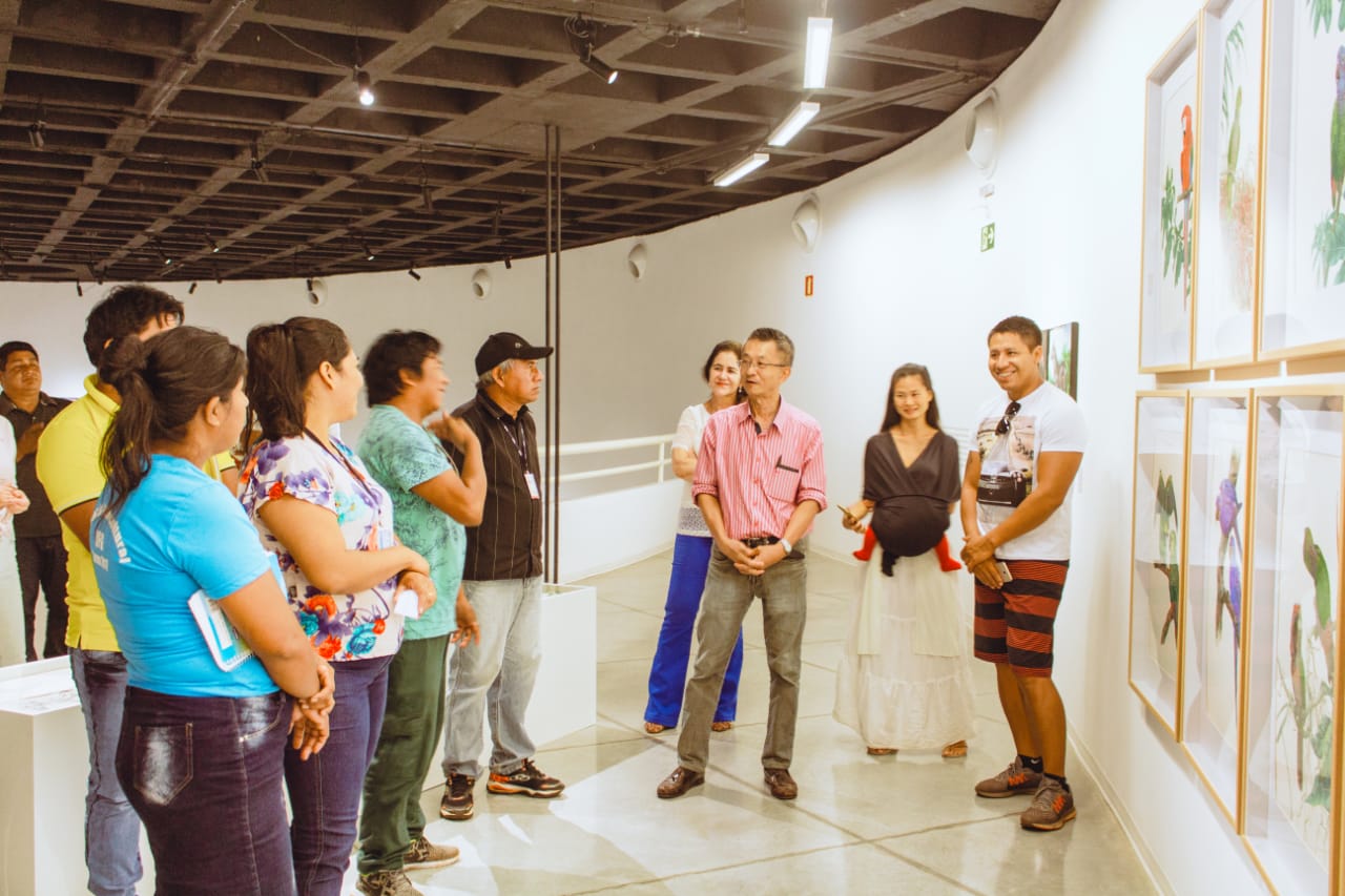 Tai conversando com os estudantes indígenas da Universidade Federal de Goiás (UFG) que foram visitar sua exposição. Estavam em frente às ilustrações científicas de araras e periquitos.
A filha de Tai, Lian, está com um bebê no colo. Ela é ativista dos direitos indígenas e por acaso foi ao encontro. Ao final, convidaram-a para visitá-los na UFG.
Exposição no Brasil com as mesmas obras que foram expostas na Expo China 2018. Foram exibidas no Museu de Arte Contemporânea de Goiânia (GO).
Entre as obras, apresentou desenhos, gravuras, instalações e esculturas também.