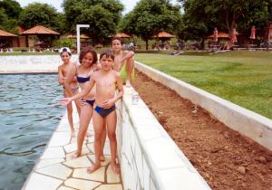 Suelen, filha de Maria Amélia, junto aos primos Henrique e Marcelo no clube, em trajes de banho, ao lado da piscina.