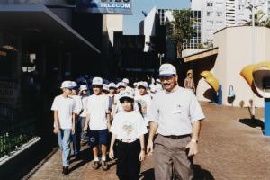 Durval está caminhando com um grupo de crianças em frente à CTBC, num calçadão. A primeira menina, à frente, é filha de Durval. Foi ela quem pediu para fazerem a visita. As crianças queriam conhecer o funcionamento de telefonia fixa e móvel.