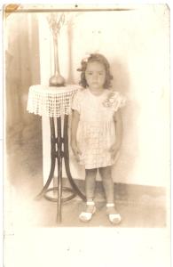Com 5 aninhos, com vestido cor de rosa com flores e lacinho nos cabelos ruivos, a pequena Bernadette se prepara para a festa, mas antes, uma foto na casa de sua avó perto da mesinha de madeira.