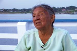 Mulher sentada em um píer. Há o oceano, árvores e casas ao fundo.