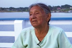 Mulher sentada em um píer. Há o oceano, árvores e casas ao fundo.