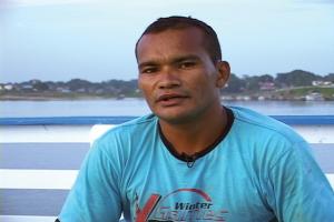 Homem sentado em um píer. Há o oceano; árvores e casas ao fundo.