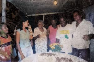 A fotografia registra Adriana em sua juventude, acompanhada da sogra, Dona Martinha, e do marido Sebastião, conhecido como Zumbi, durante uma celebração com amigos na Vila Esperança. A cena revela a força dos laços familiares e comunitários, marcando um momento de convivência festiva.