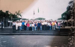 Adriana conta que a comunidade lutou contra a falta de água e luz e que a maior conquista foi a urbanização, com 15 km de cerca para conter invasões e garantir infraestrutura para a comunidade de Vila Esperança. Apesar das dificuldades, a eleição de representantes locais trouxe esperança de avanços reais. A imagem registra um momento de reunião entre Zumbi, seu esposo e líder da comunidade, e as outras lideranças comunitárias e do poder público no pátio da Prefeitura de Cubatão.