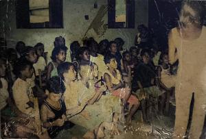 Anderson, conhecido como Andinho, aparece em destaque no centro da imagem, sentado e com olhar atento, acompanhando uma aula. De compleição magra, sua postura transmite concentração e interesse. A cena integra uma atividade de formação vinculada a um programa social realizado na década de 1990, na Vila Esperança, em Cubatão. O espaço pertence ao líder comunitário Zumbi, figura central em diversas ações educativas e culturais da região. Embora o conteúdo da aula não esteja identificado, o registro sugere um momento de escuta e aprendizado coletivo.