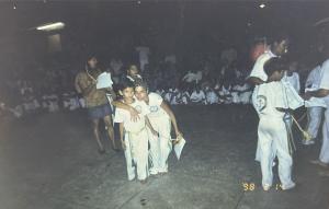Registro do batizado de capoeira do Grupo Brasil/Suíça, no antigo Bingo Vegas, em Cubatão. Patrícia aparece ao lado de seu afilhado Meison, em um momento ritual de iniciação na capoeira. A fotografia também se relaciona à trajetória de Patrícia como capoeirista, marcada pela formação, pelo reconhecimento e pela conquista de diploma, reafirmando a capoeira como espaço de aprendizado, pertencimento e continuidade cultural.