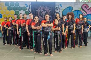 Patrícia aparece acompanhada de suas alunas durante um evento de capoeira. À sua esquerda estão Ana Clara, Isa, Letícia, Lívia, Paloma e Beatriz; à direita, Larissa, Ana, Nathália, Samira, Rachel, Mylla e Beatriz. A fotografia registra o grupo reunido, destacando a presença feminina na capoeira e a relação entre mestra e alunas em um contexto coletivo e formativo.