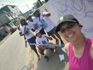 Patrícia aparece ao lado de integrantes do projeto da Rumo durante a ação de grafitar o muro da Vila Esperança. A imagem registra o artista Marco Tuim em atividade pelo Projeto da Linha Pra Frente, enquanto o grupo participa do processo de colorir e transformar a paisagem do bairro.