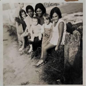 No centro da imagem está Dona Cícera, ainda jovem, em um momento de convivência com amigos e crianças, datado entre o final dos anos 1960 e início dos anos 1970. Ela aparece com um bebê no colo, cercada por outras crianças, em uma cena que transmite afeto e coletividade. O registro fotográfico, raro para o período e contexto social, representa não apenas um instante de encontro, mas também a valorização da memória e laços comunitários.