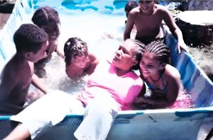 Cícera aparece ao centro da imagem, dentro da piscina, acompanhada por seus netos em um momento de descontração familiar. A cena revela o vínculo afetivo entre gerações e a importância dos espaços de lazer compartilhado no cotidiano da família. O registro carrega a leveza de um encontro íntimo, marcado pela presença ativa de Cícera junto às crianças, reforçando sua centralidade como figura cuidadora e agregadora no núcleo familiar.