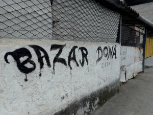 Localizado em frente à casa de Dona Cícera, o Bazar da Cissa foi sua principal fonte de renda nos últimos anos antes de passar a utilizar cadeira de rodas. Por muito tempo, ela manteve o pequeno comércio ativo, cuja fachada ainda exibe o nome do empreendimento, preservando a memória de sua dedicação ao trabalho. Parte das roupas que compunham o estoque ainda estão com ela, como testemunho material dessa trajetória. A imagem retrata um dos aspectos centrais de sua vida: o envolvimento com o comércio local. Nos anos 1990, Dona Cícera também foi proprietária, junto ao então marido Tigrão, do Bar do Forró, um ponto de encontro muito conhecido na Vila Esperança, em Cubatão, que marcou a vida cultural da comunidade.