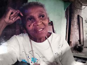 A imagem retrata Dona Martinha, mãe de Zumbi, em um momento introspectivo. Moradora da Ilha Bela desde o início dos anos 1980, ela foi reconhecida como uma importante líder comunitária e como forte inspiração para o trabalho de seu filho, Sebastião, o Zumbi, que segue atuante como liderança local. A fotografia valoriza a presença feminina nas lutas do território e a influência das mulheres na construção da vida comunitária.